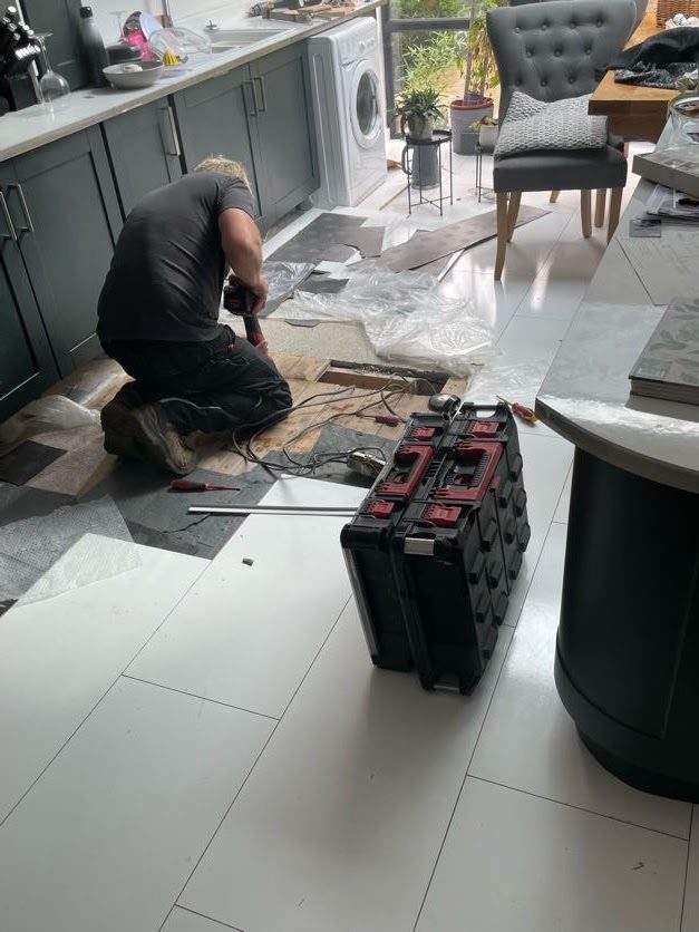 Kev kneeling at lifted floorboards in a kitchen, tool cases beside him
